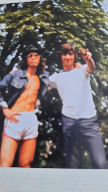 Zwei Männer im Freien, einer trägt ein Muskelshirt und der andere ein T-Shirt, beide lächeln.