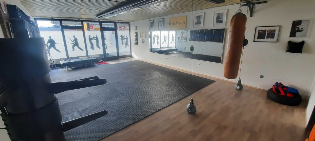 Leerer Trainingsraum mit Boxsack, großem Mattenbereich und Fenstern im Hintergrund.